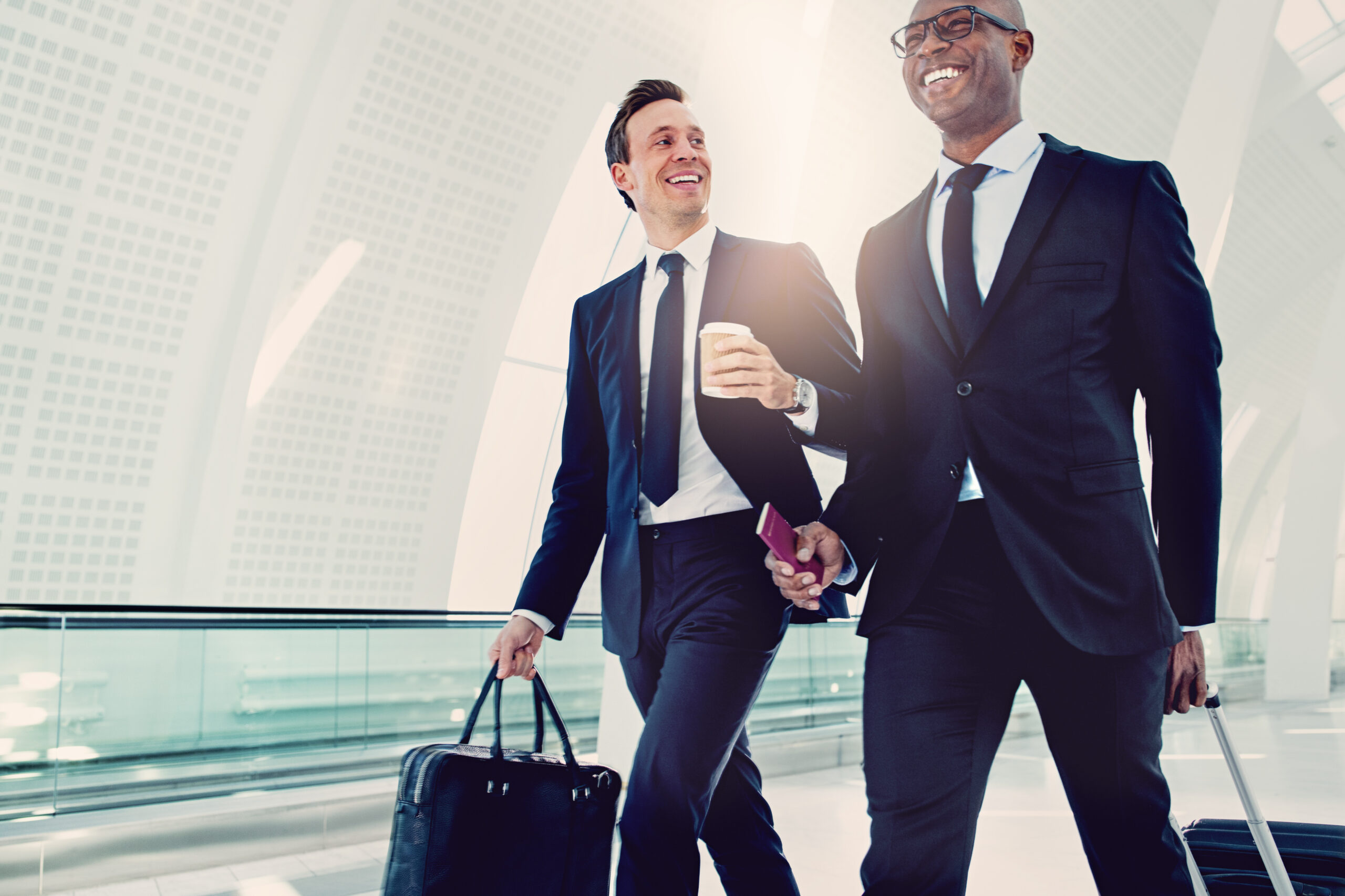 Two businessman in suits smiling and talking together while walking with their baggage down a corridor in a bright modern airport
